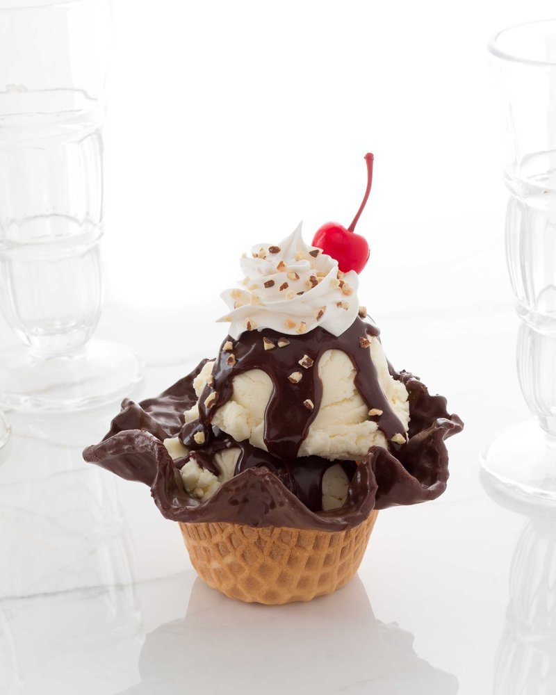 Ice cream sundae in waffle cone bowl with chocolate sauce, whipped cream, and cherry.