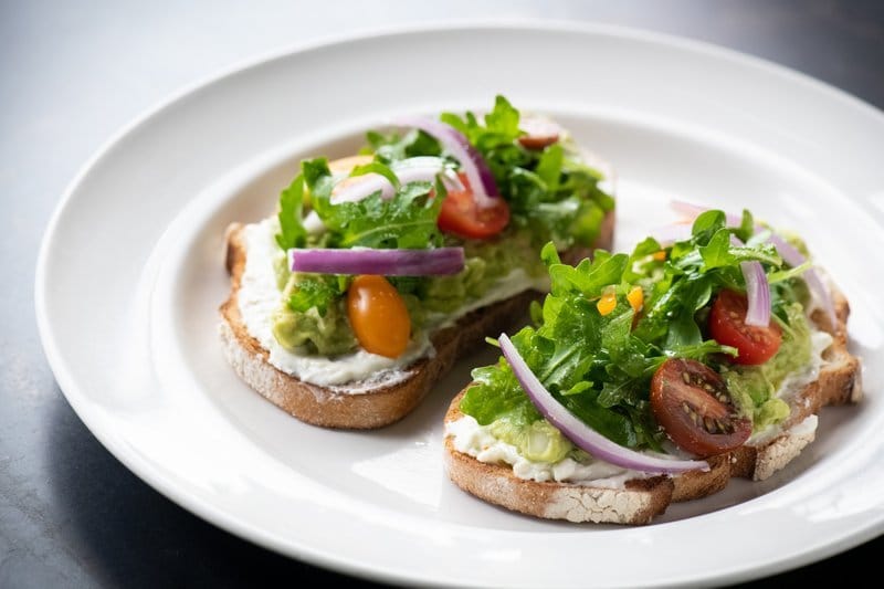 Avocado toast with greens, tomato, and onion on a white plate.