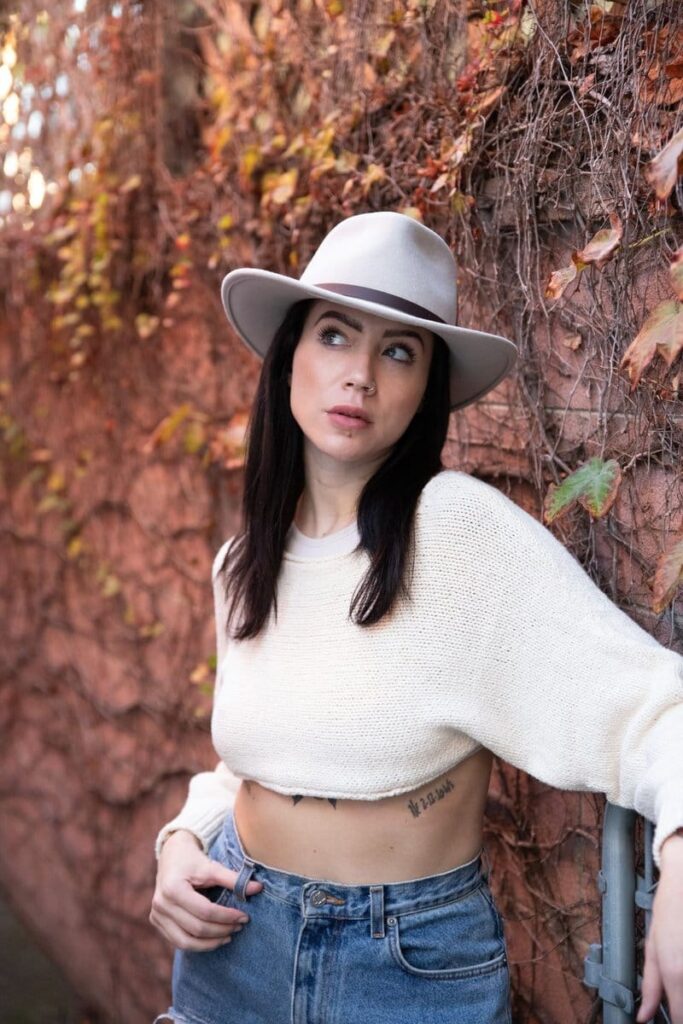 Woman in hat and sweater posing against ivy wall.