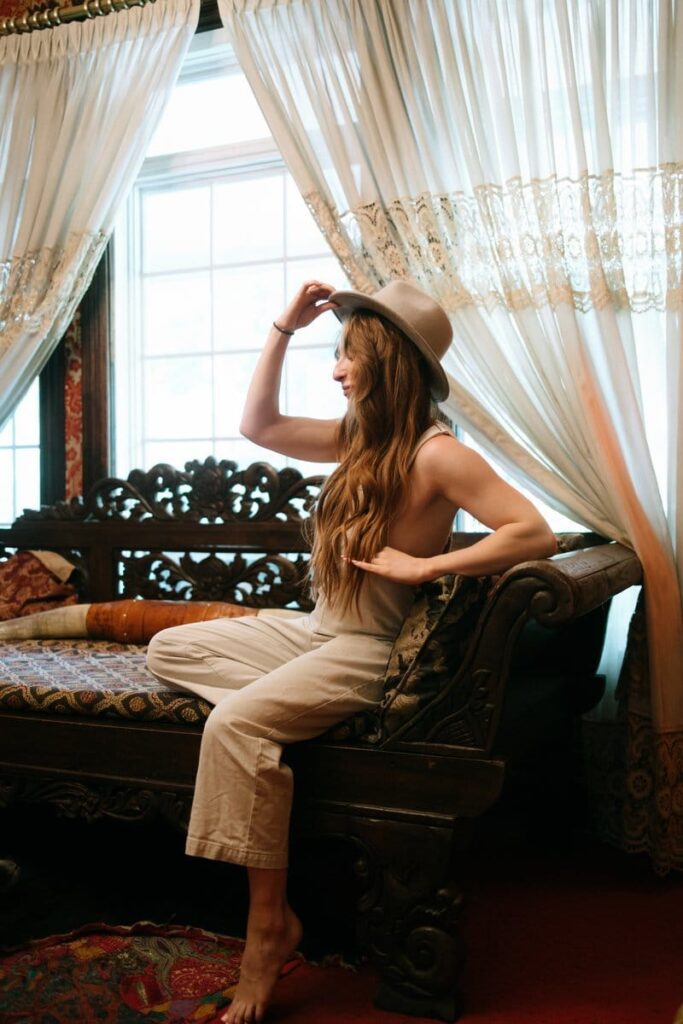 Woman with hat seated on ornate bench near window.