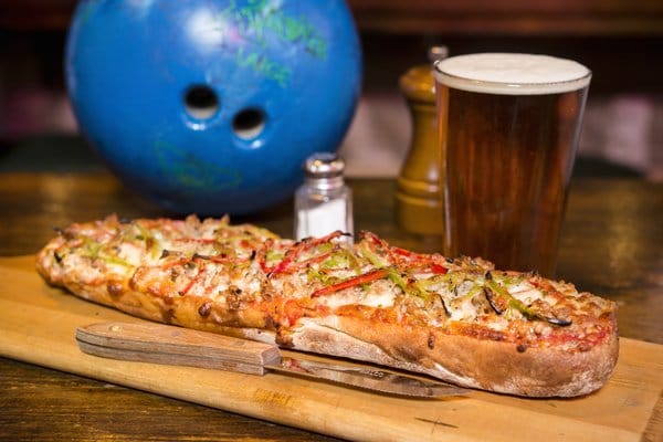 Long pizza on wooden board, bowling ball, beer.