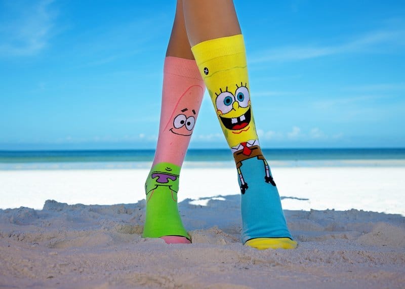 Legs wearing SpongeBob and Patrick socks on a beach.